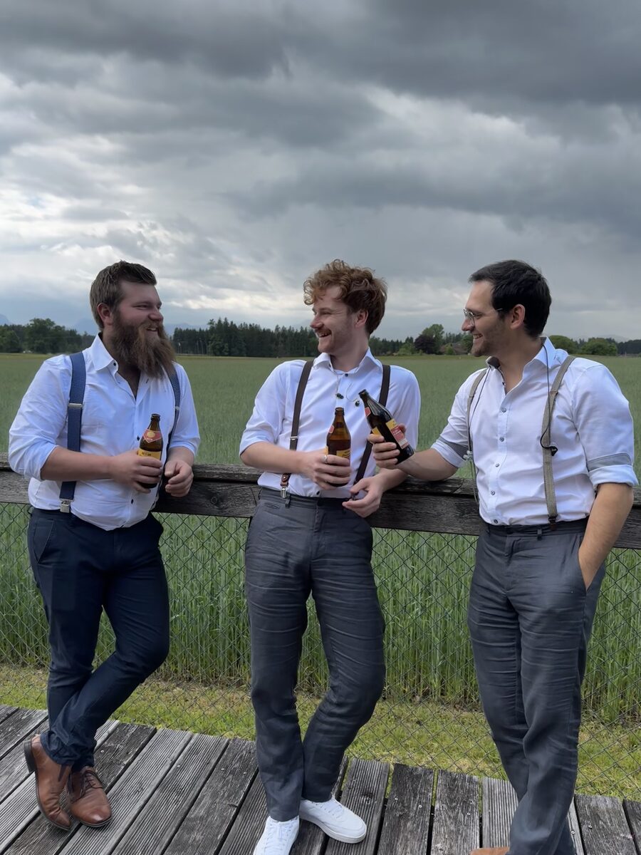 Drei Musiker von Boazn Royal stehen lachend mit Getränken vor einem Feld